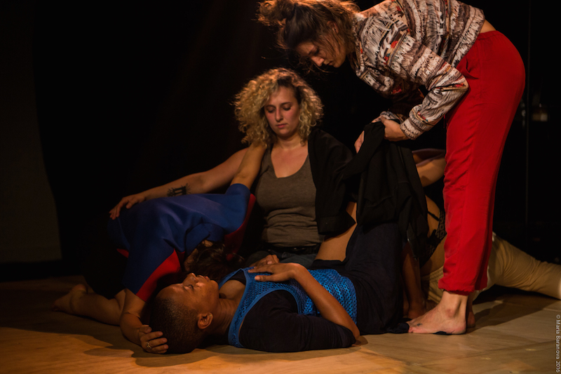 Jennifer Harge lays on her back while another dancer curls up next to her. Her face is hidden. Nadia Tykulsker knees besides them while EmmaGrace Skove Epps stands over them.